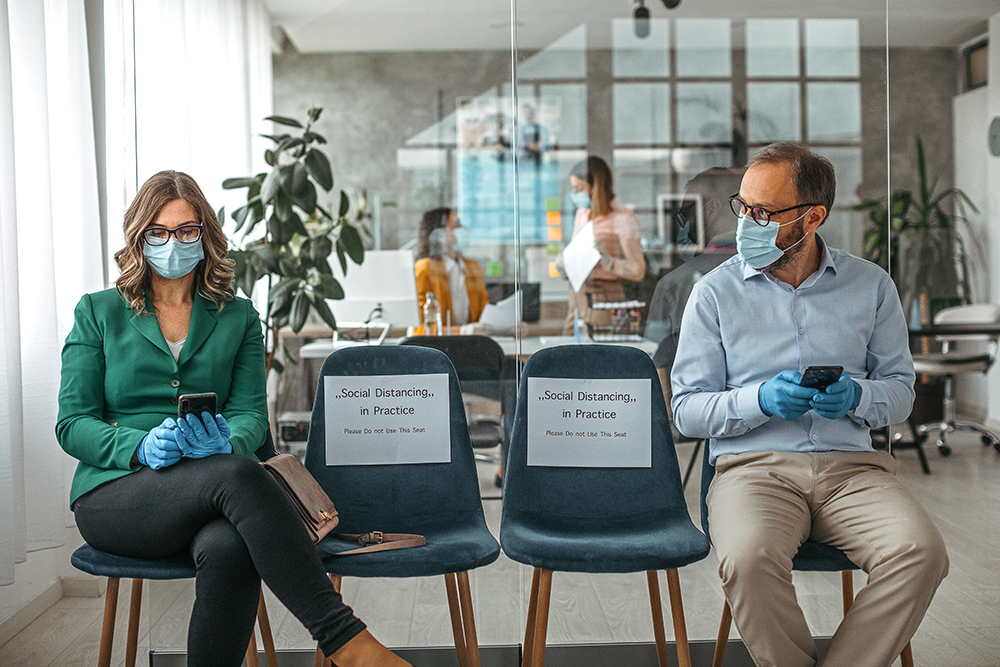 Customers using mobile phone wearing protective masks and gloves sitting on office chair while maintaining social distance to protect from COVID-19