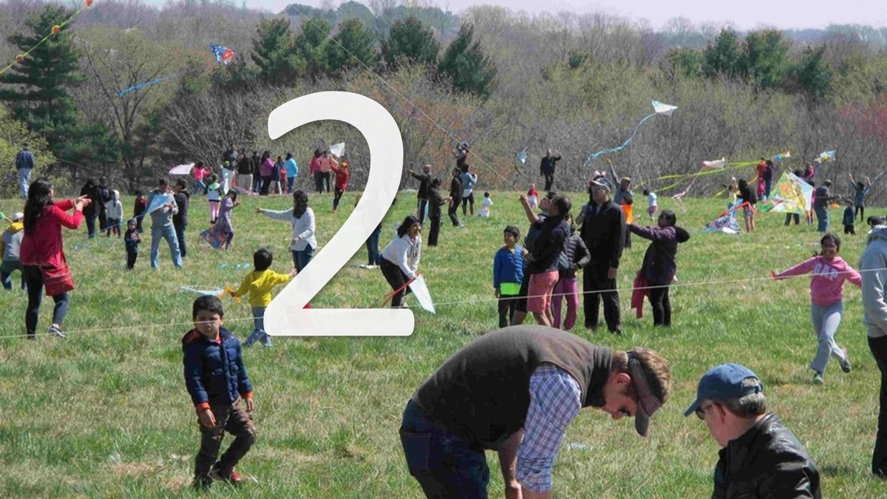 families with young children flying kites together in an open grassy field with trees in the background and a number two in front of the picture