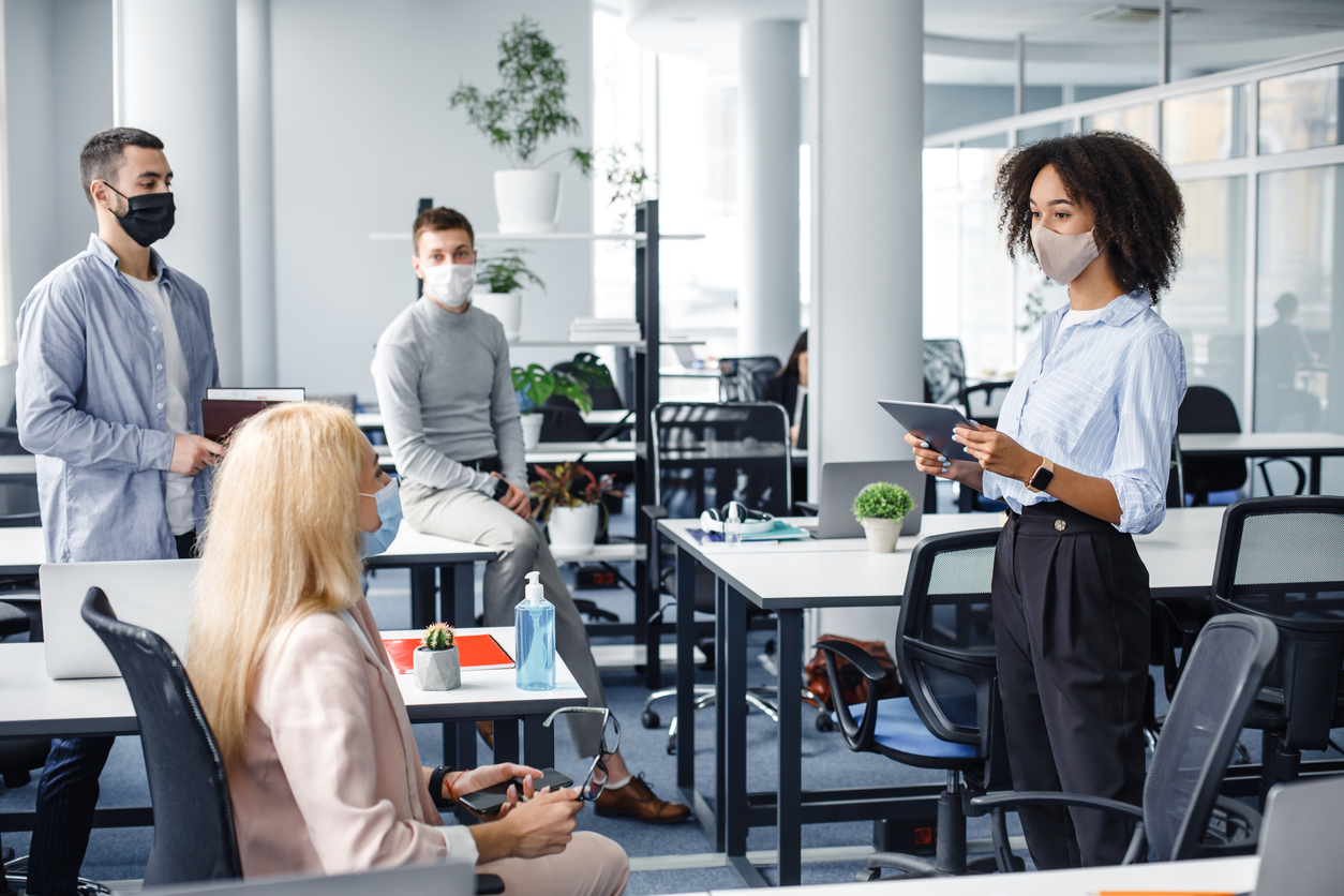 Corporate meeting and group work in modern company in office interior. African american woman manager in protective mask holding tablet, talking to workers keeping social distance during epidemic