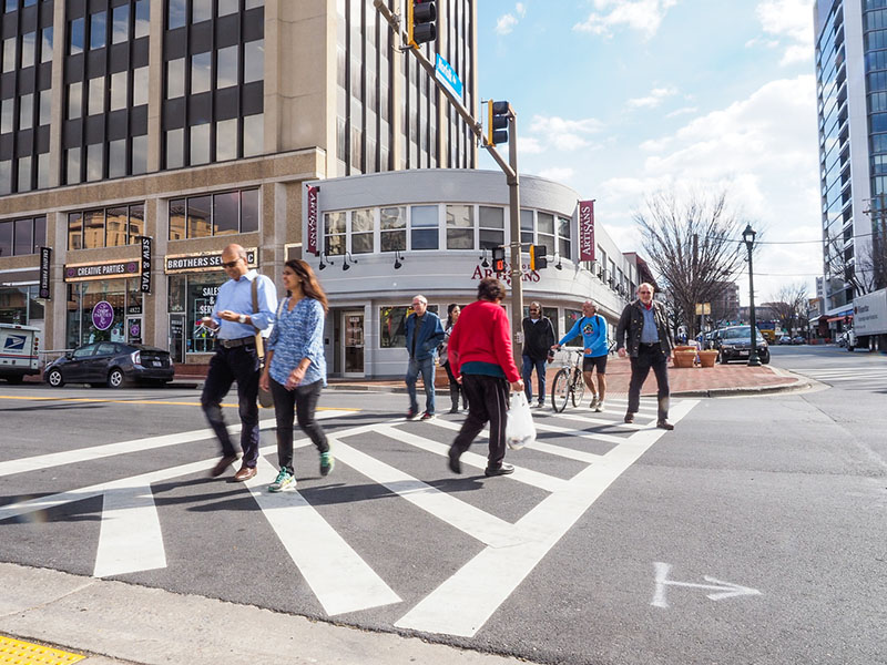 people are crossing a marked crosswalk in Bethesda, including a seven people walking towards the photographer (including one man walking a bicycle) and a woman walking away from the photographer. 