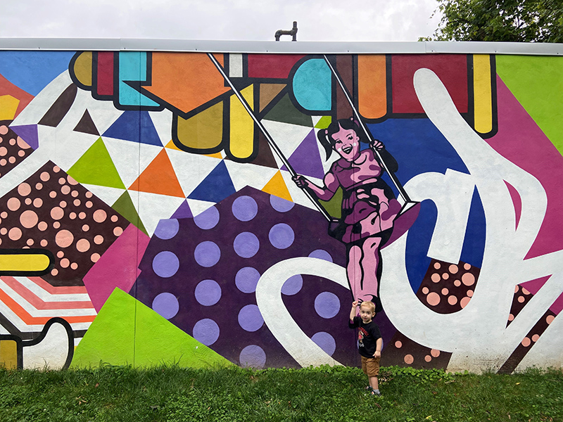 toddler standing in front of bright colored mural featuring a girl on a swing at Dewey Local Park