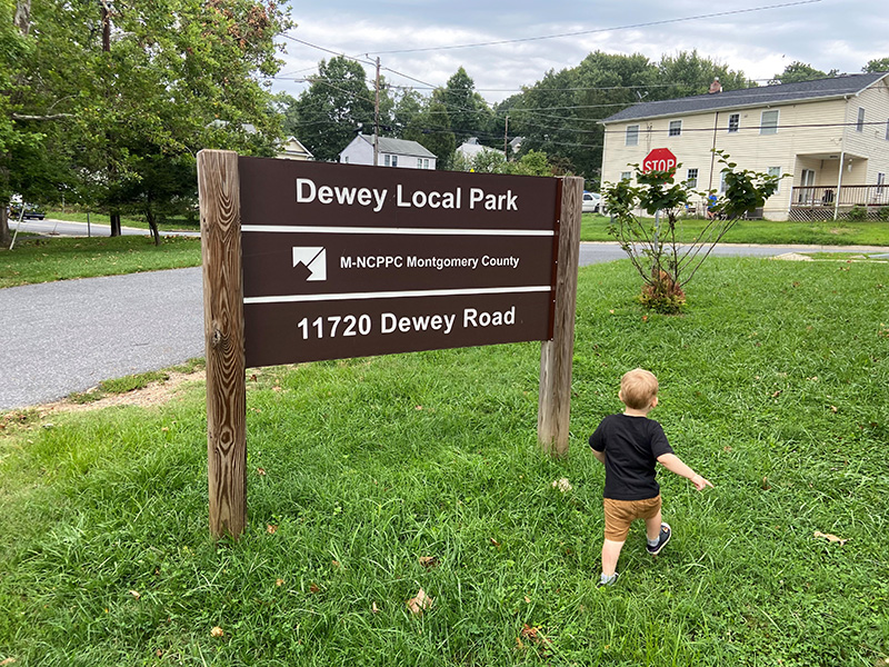 child walking past Dewey Local Park wooden sign 