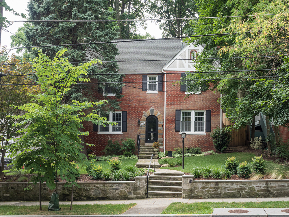 A two-story brick house with a symmetric design, featuring a central arched doorway flanked by two windows on each side and three dormer windows on the roof. Landscaped with various shrubs and trees, the house is approached by a series of steps leading to the main entrance.