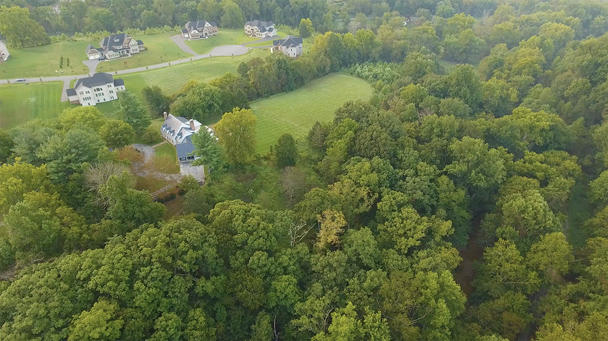 Aerial view of a residential neighborhood surrounded by dense green trees. Several houses with large yards are visible, set along a curved road. The landscape includes a mix of open grassy areas and wooded sections.