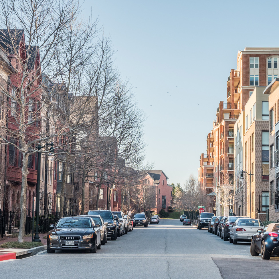 A quiet city street with parked cars on both sides. Leafless trees line the sidewalks next to a mix of brick and modern buildings under a clear blue sky.