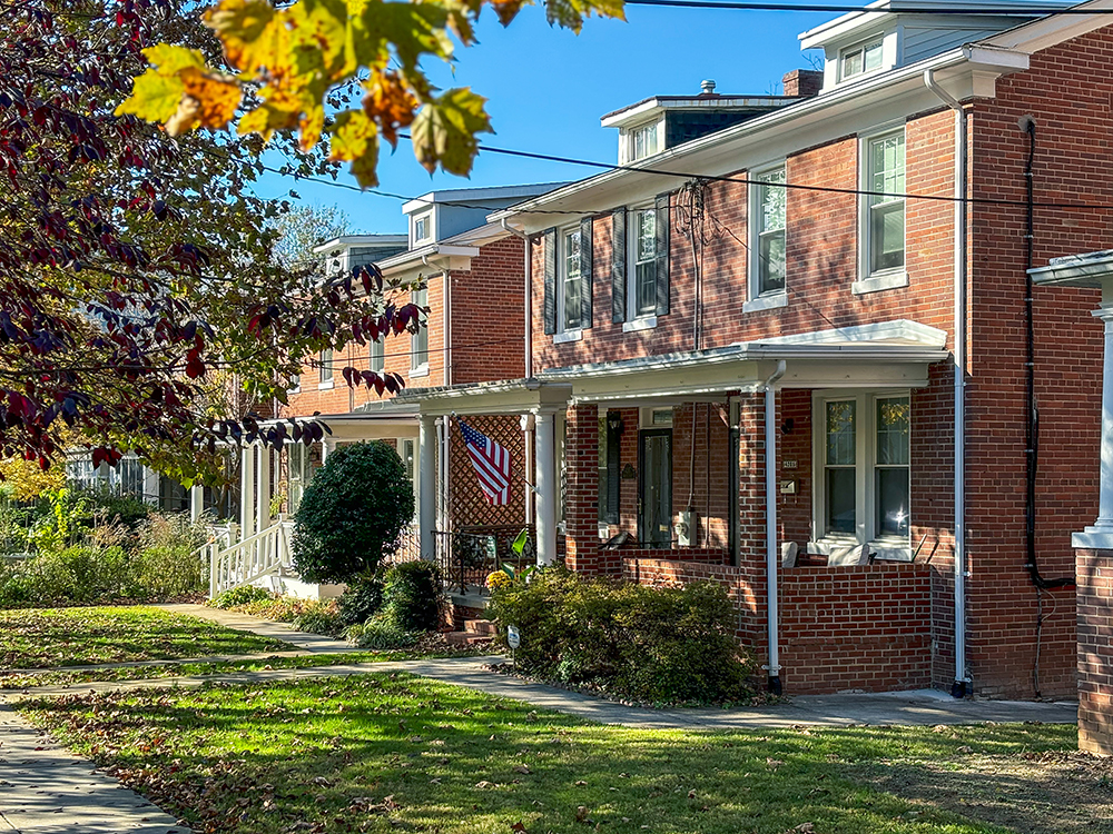 A row of red brick houses with white trim on a sunny day. An American flag hangs from one porch. Green lawns and a tree with autumn leaves are visible in the foreground.