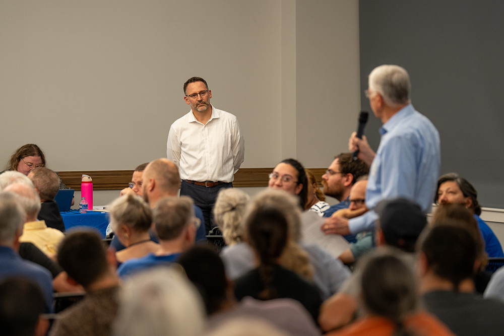 Jason K Sartori stands in front of a seated audience, listening to someone holding a microphone during a meeting. Papers and a pink water bottle are on a table in the background.