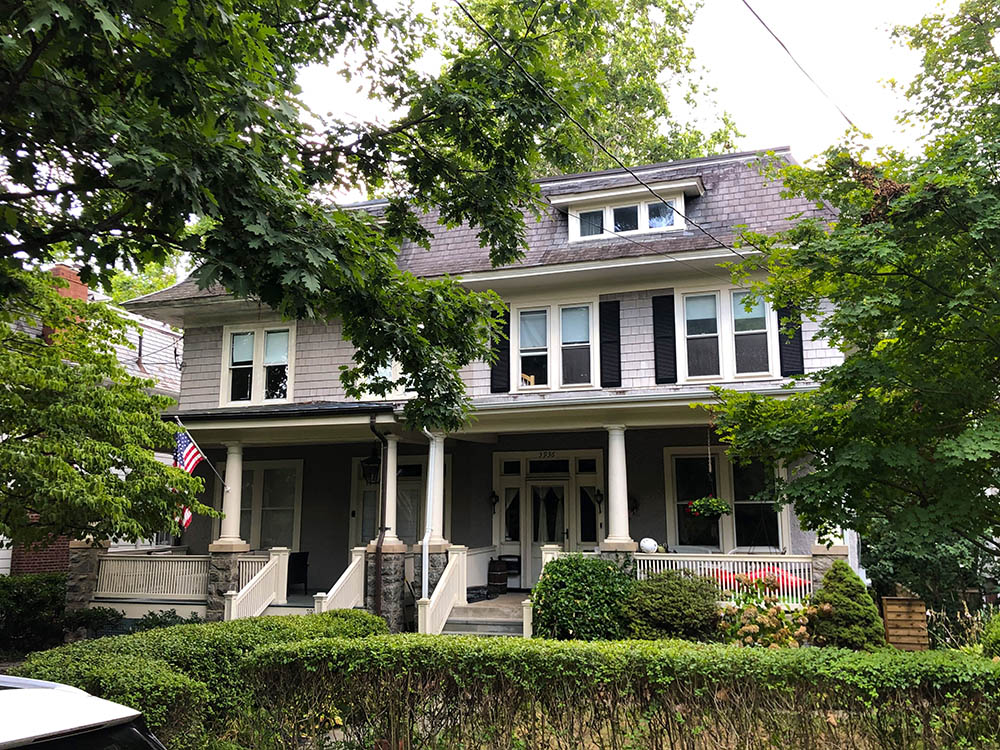 A large, two-story house with a porch, surrounded by lush green trees and bushes. An American flag hangs by the entrance. The exterior is shaded, with a mix of gray siding and white trim.