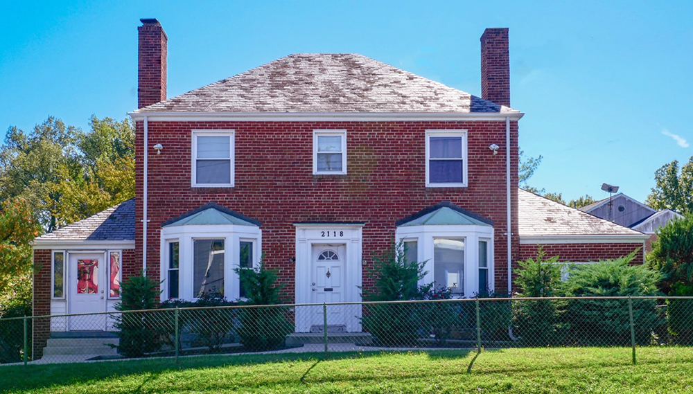 A two-story brick house with a symmetrical design, featuring two chimneys and two bay windows. The front has a white door with the number "2118" above it. A green lawn and a chain-link fence are in the foreground, with trees in the background.