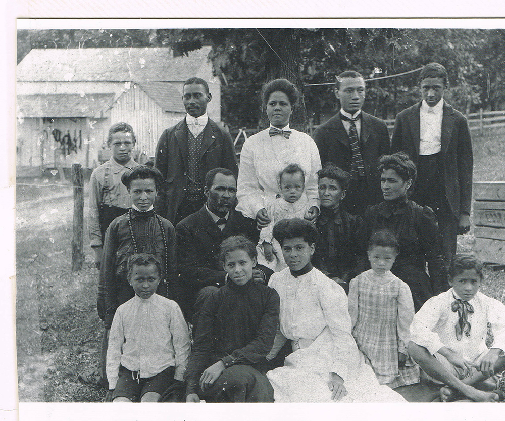 A vintage black and white photo of 14 people, including men, women, and children, posed outdoors. Most are seated or standing on grass near a wooden building. The group wears formal attire typical of the late 19th or early 20th century.