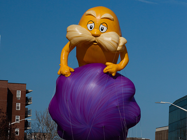 A large, colorful inflatable balloon featuring a yellow character with a bushy mustache sitting atop a purple sphere. The balloon is set against a clear blue sky and is surrounded by buildings and streetlights.