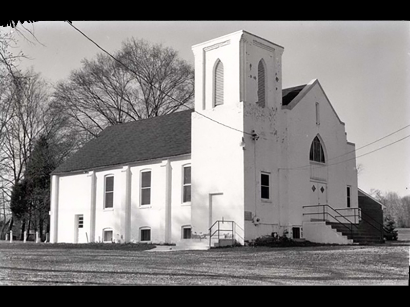 A black-and-white photo of a small, white church with a steeple. The building has arched windows and a set of stairs leading to the entrance. It's surrounded by bare trees under an overcast sky.