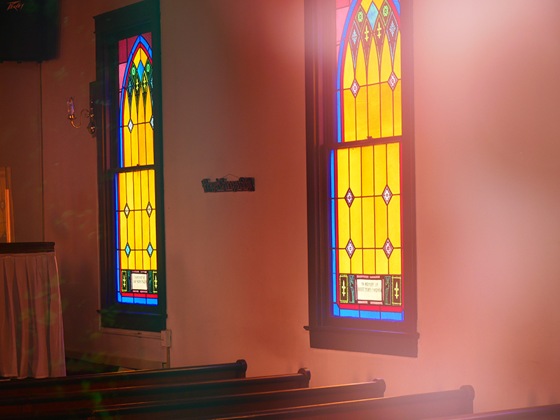 Interior of a church with sunlight streaming through two colorful stained glass windows, casting vibrant colors on the wooden pews below. The windows feature geometric and floral designs in yellow, blue, and red hues.