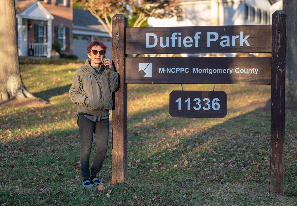 A person wearing sunglasses, a gray jacket, leggings, and sandals is standing next to a large wooden sign that reads "Dufief Park, M-NCPPC Montgomery County, 11336." There are houses and trees in the background.