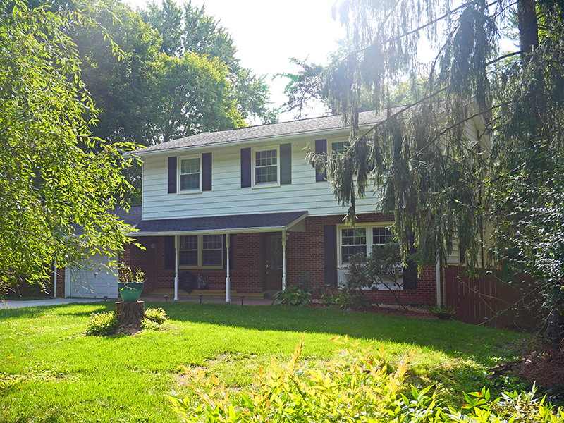 A two-story house with a mix of brick and white siding exterior, dark shutters, and a covered porch. The front yard is lush with grass, bushes, and a large tree. Sunlight filters through the trees, casting dappled shadows on the lawn.