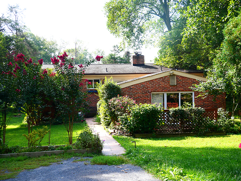 A charming red brick house with a sloped roof, surrounded by lush greenery and blooming bushes. A narrow stone path leads to the front, bordered by vibrant flowers and shrubs under a clear, sunny sky.