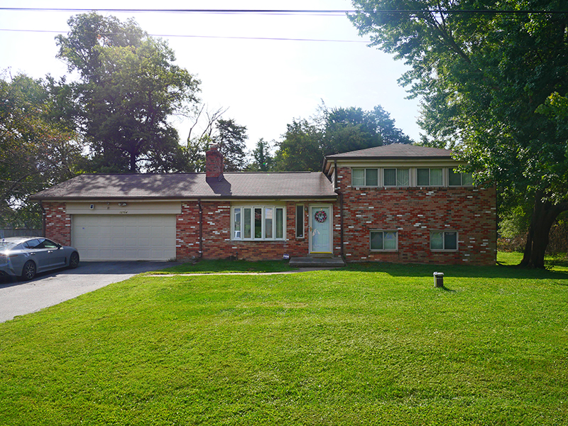 Single-story brick house with a two-car garage, front lawn, and driveway. The house features a large window with white shutters and a small porch with a wreath. Trees surround the property, and a car is parked in the driveway.