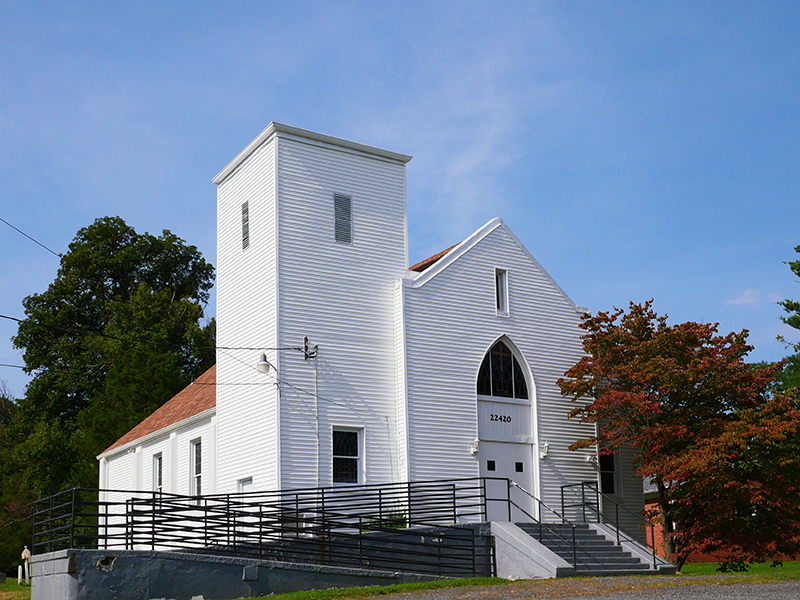 A white wooden church with a tall square tower and brown roof stands under a clear blue sky. It has a large arched window and a ramp leading to the front entrance. A tree with red leaves is on the right.