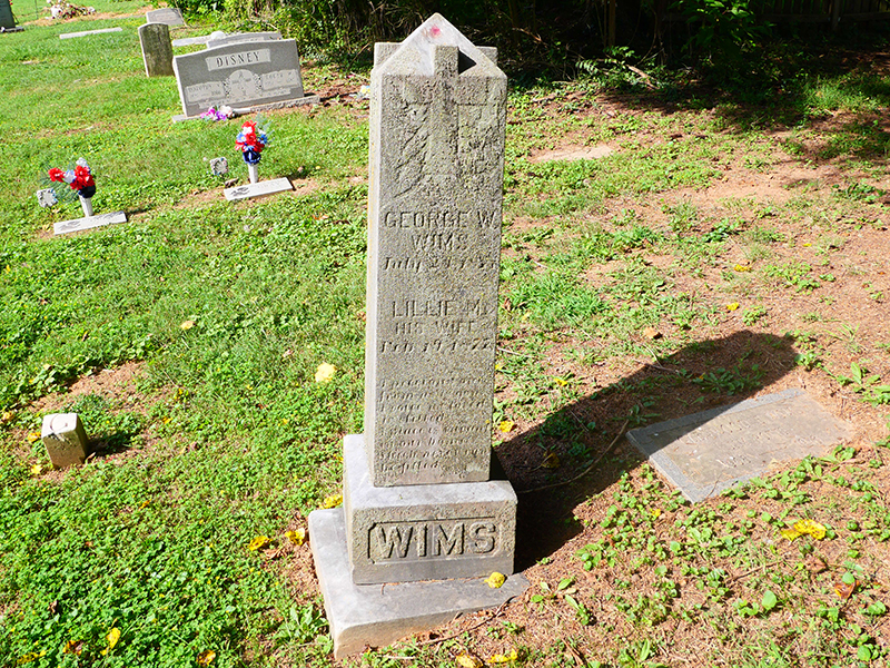 An old, weathered tombstone surrounded by grass and other graves. It is inscribed with the names George W. Wims and Lillie M. Wims. The area is scattered with flowers, and another gravestone is visible in the background.