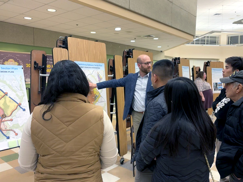 A man in a suit gestures towards a large map on a display board, explaining to a group of six people in casual attire. The setting appears to be a community meeting or presentation in a room with tiled floors and overhead lights.