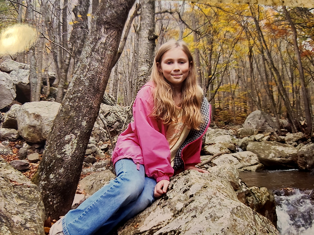 Girl in a pink jacket and jeans sitting on a rock in a forest by a stream. Trees with yellow and brown leaves surround her, suggesting autumn. She looks at the camera, with a backdrop of rocks and flowing water.