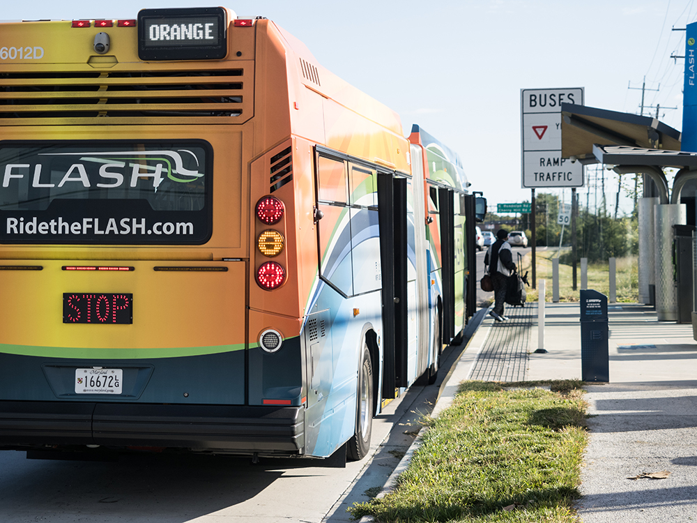 A colorful city bus labeled "ORANGE" is stopped at a bus station. People are boarding near signs for buses and ramp traffic. The bus displays "STOP" and the website "RidetheFLASH.com" on the back.