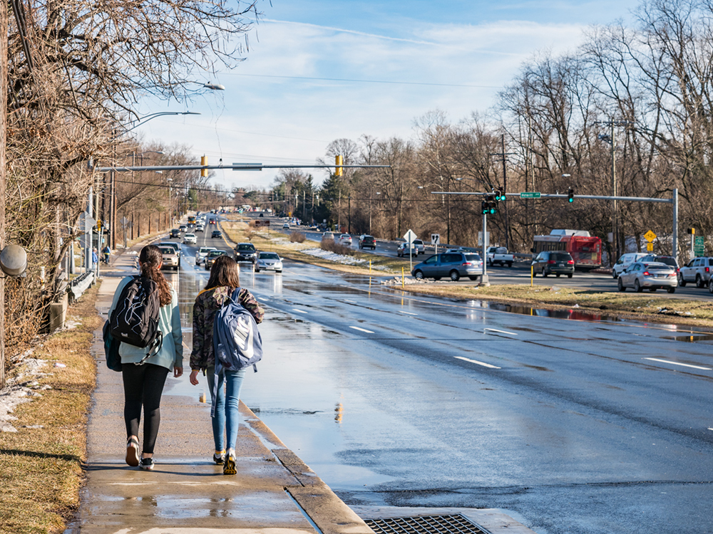 Two people with backpacks walk on a sidewalk beside a busy road with puddles, traffic lights, and cars. Trees without leaves line both sides of the street under a blue sky with clouds.