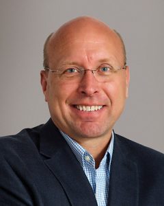 A smiling middle-aged man with glasses, a bald head, and light skin, wearing a dark blazer over a blue checkered shirt, posed against a plain light background.