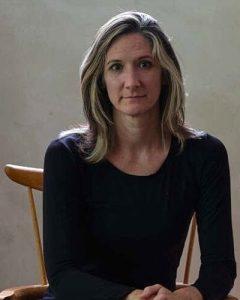 A woman with shoulder-length, wavy, light brown hair sits on a wooden chair, wearing a long-sleeved black top and looking calmly at the camera. The background is plain and softly lit.