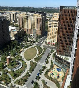 Aerial view of a cityscape with tall buildings, green park areas, walking paths, a circular lawn, a small playground with colorful equipment, and tree-lined streets under a clear sky.