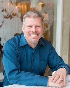A smiling man with short gray hair and a beard, wearing a teal button-up shirt, sits at a table with his hands folded. The background features a glass wall with a white, abstract dotted design.