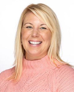 A smiling woman with straight blonde hair wearing a light pink, cable-knit turtleneck sweater, posed against a plain white background.