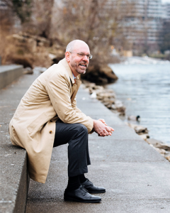 A man wearing glasses, a beige trench coat, and dark pants sits on a concrete ledge by a river, smiling and looking into the distance. Trees and buildings are visible in the background.