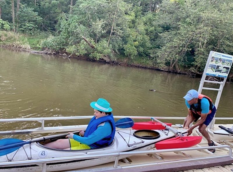 Two people prepare a white kayak on a dock by a river; one person sits inside wearing a turquoise hat and blue vest, while the other adjusts the kayak’s front, surrounded by trees in the background.