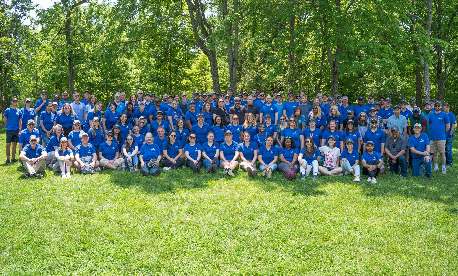 A large group of people wearing matching blue shirts pose together for a group photo outdoors on a grassy area with trees in the background.