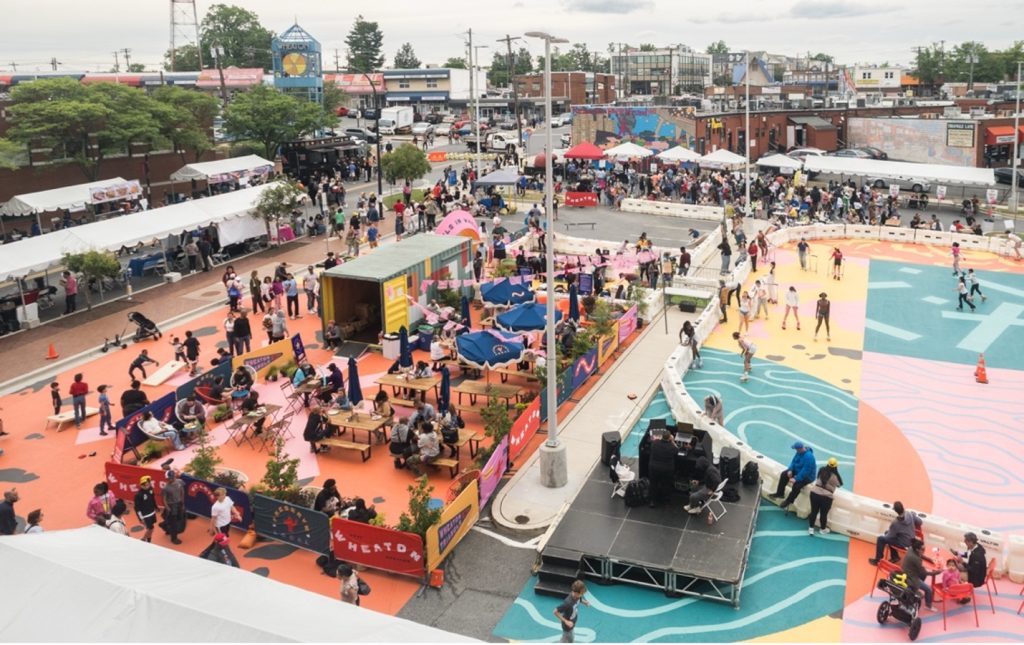 A vibrant outdoor community event with colorful painted ground, vendor tents, tables, people walking and sitting, and a stage with equipment; murals decorate nearby walls under a cloudy sky.
