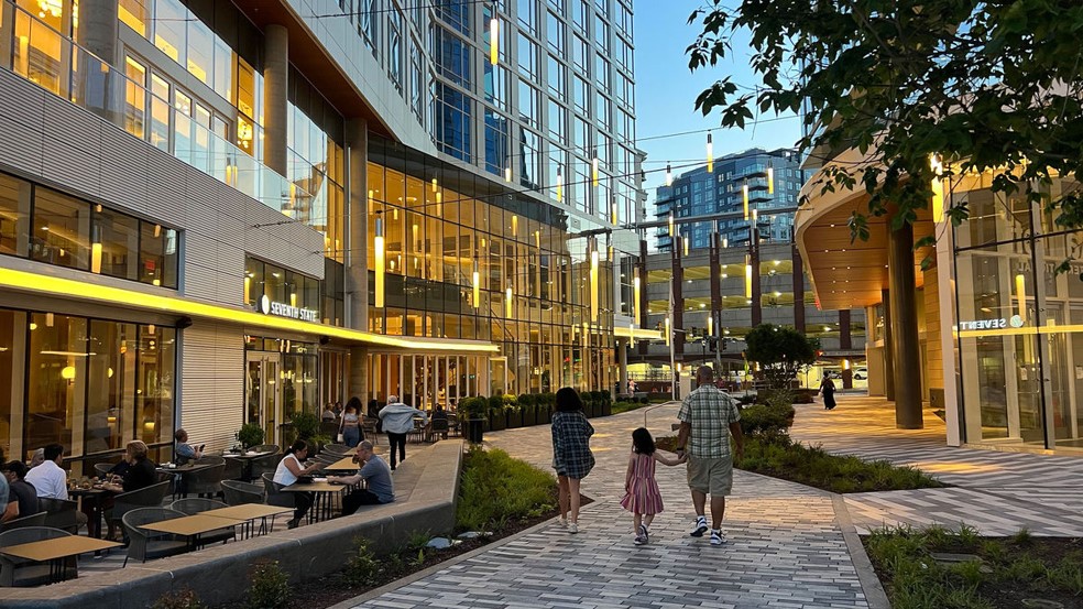 A family walks along a modern outdoor plaza surrounded by glass buildings and lit-up restaurants at dusk, while people dine at outdoor tables. Trees and greenery line the walkway.