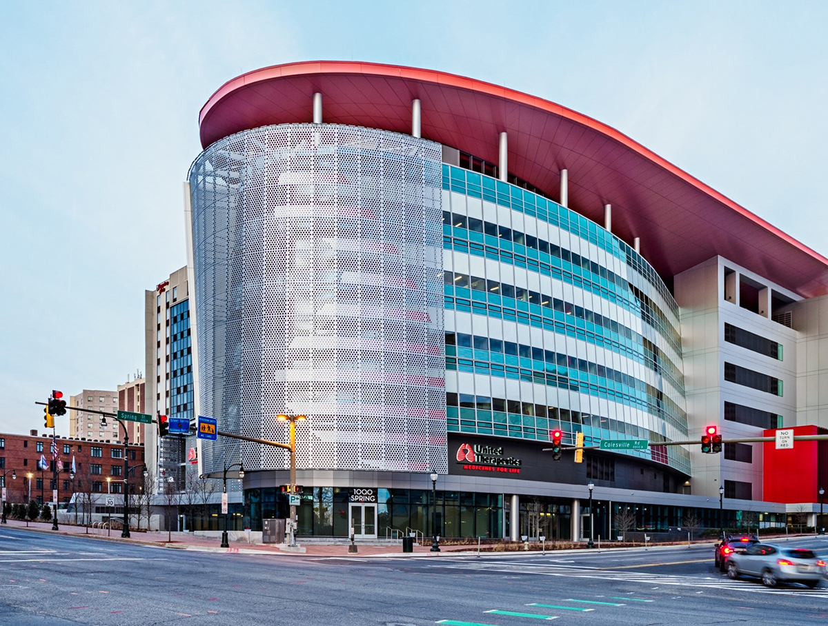 An oval-shaped multi-story building with a red roof situated on a corner lot in an urban setting.