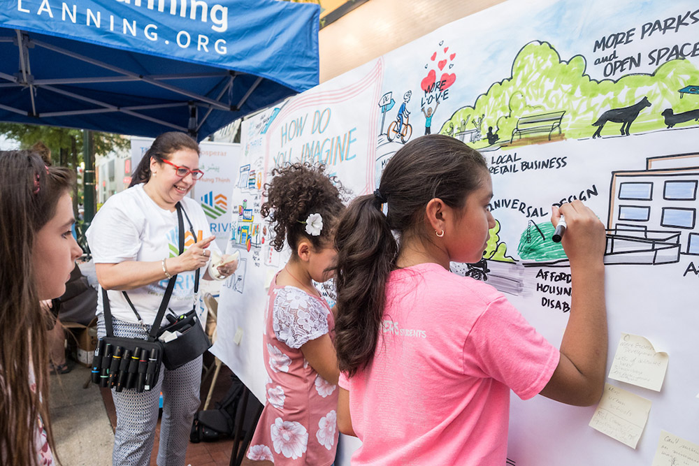 Children and an adult draw and write on a large community mural under a blue tent, illustrating ideas for urban planning such as parks, green spaces, and local businesses.