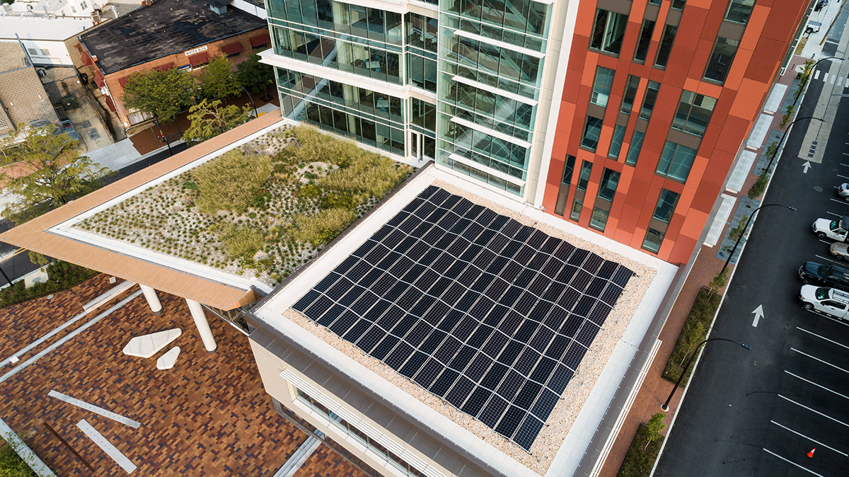Aerial view of a modern building with a green rooftop garden and solar panels, surrounded by city streets and parked cars.