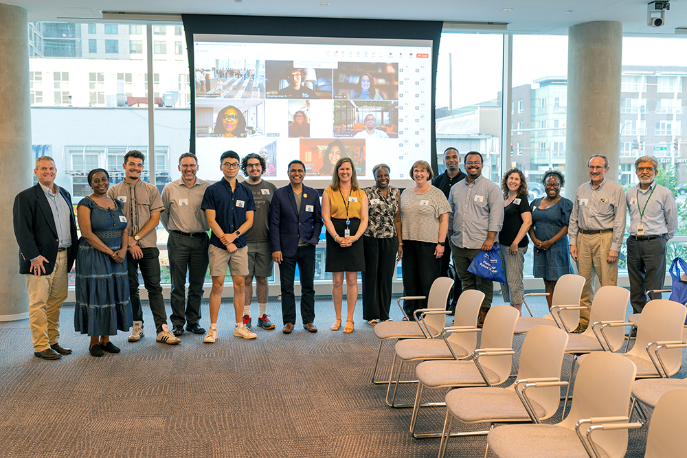 A group of fifteen people stand in a row, smiling at the camera in a modern conference room with large windows and empty chairs; a projection screen behind them shows a virtual meeting grid.