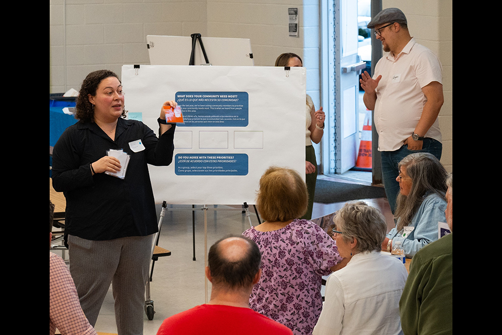 A woman stands and speaks to a seated group of people in a classroom, holding up an orange card. Behind her is a presentation board with text and diagrams, and a man stands near the open door in the background.