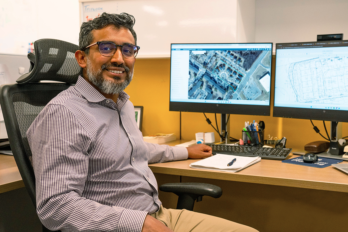 A man with glasses and a beard sits at a desk, smiling at the camera. He is in an office, with two computer monitors displaying a map and an architectural plan, along with various office supplies on the desk.