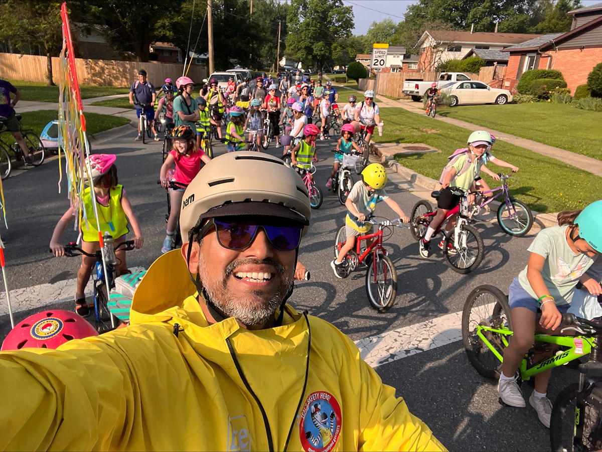 A smiling man in a yellow jacket and helmet takes a selfie in front of a group of children and adults riding bicycles down a neighborhood street on a sunny day. Many riders are wearing helmets and colorful outfits.