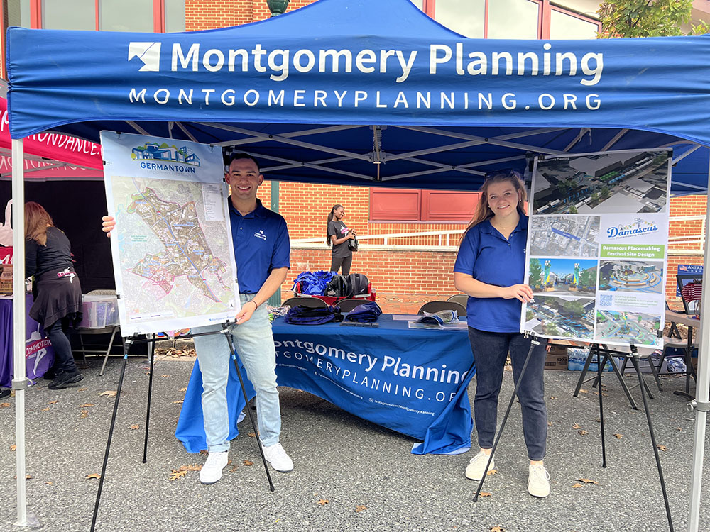 Two people stand under a Montgomery Planning tent, each holding a display board with maps and project information. A table with planning materials is behind them, and a brick building is visible in the background.