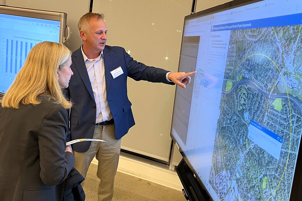 A man in a suit points at a digital map on a large touchscreen while explaining something to a woman who is holding papers. Another screen nearby displays a bar chart. They appear to be in a professional setting.