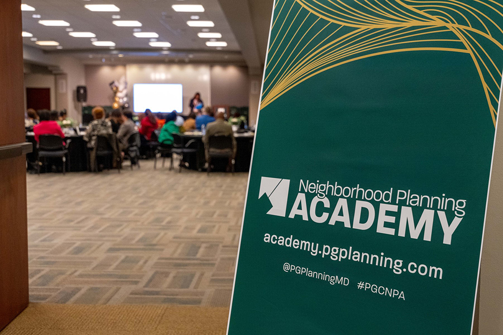 A sign for the Neighborhood Planning Academy stands in the foreground of a conference room where people sit at round tables facing a presentation screen.