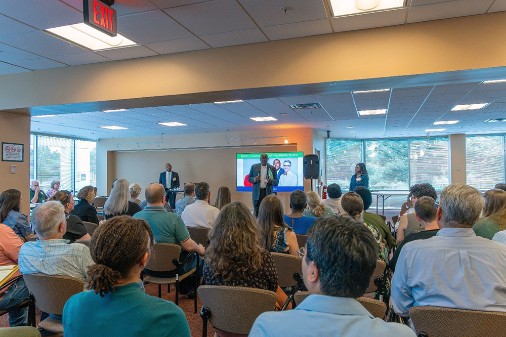A group of people sit facing a panel of speakers at the front of a well-lit room. A screen behind the speakers displays presentation slides with photos and text. Large windows line one wall.