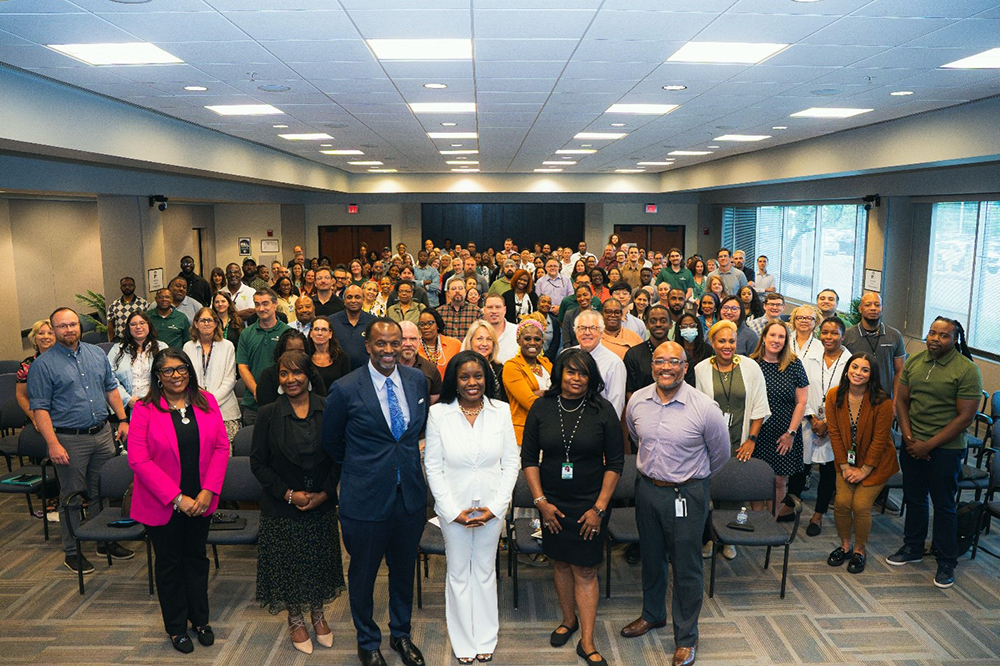 A large group of professionally dressed people stand and smile at the camera in a conference room with rows of chairs and large windows. Eight people pose at the front, while the rest gather closely behind them.