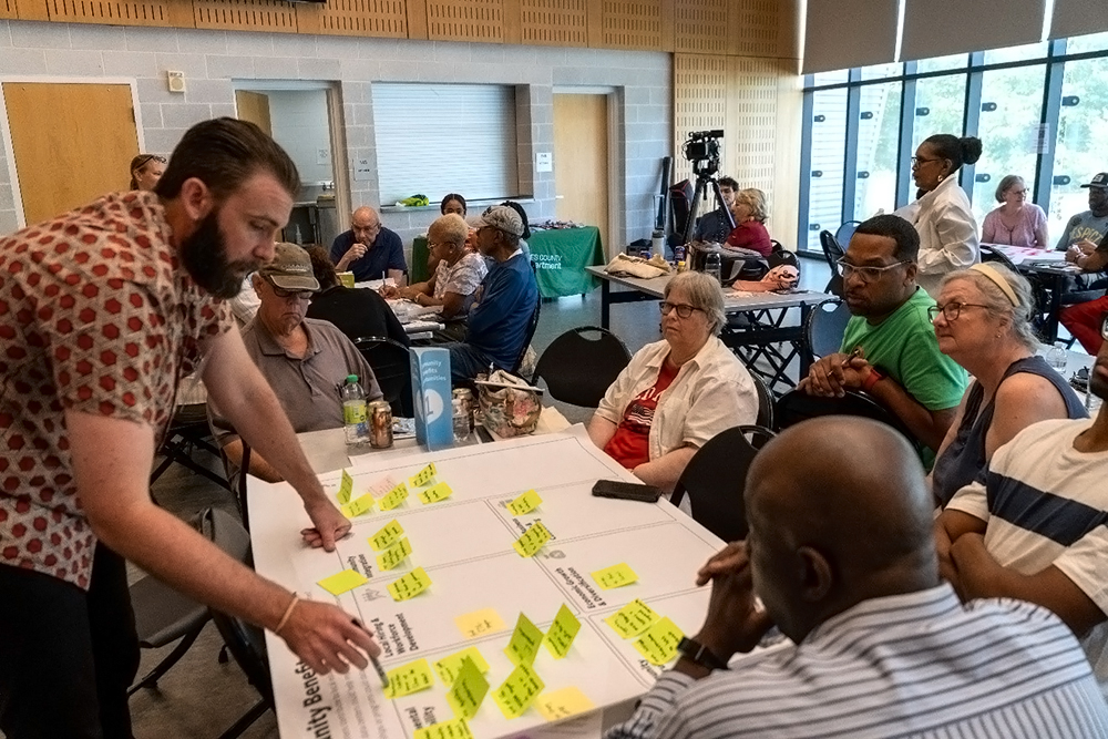 A man stands beside a table, leading a discussion with a group of adults seated around him. The table is covered with white papers and bright sticky notes. Other groups are meeting in the background of a bright, modern room.
